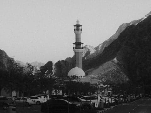 A mosque surrounded by mountains, with cars parked in the foreground and trees lining the street.