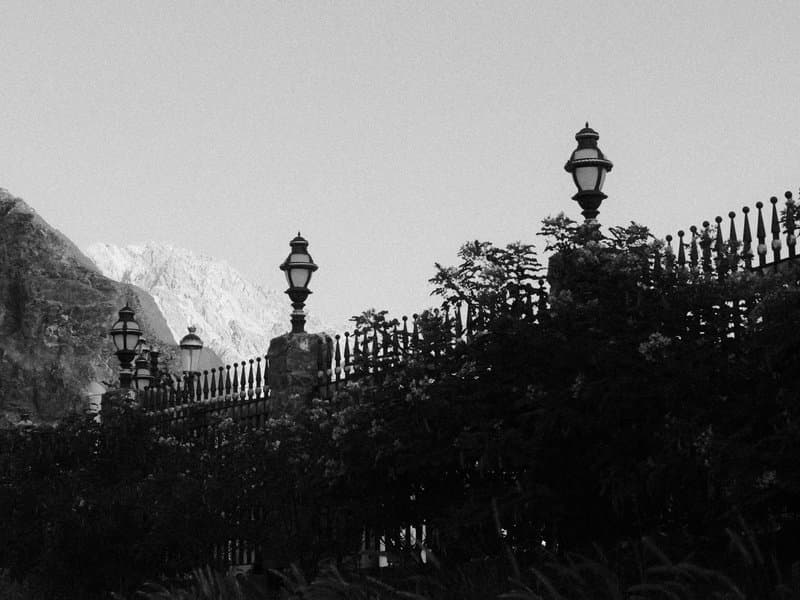 Black and white image of a decorative fence with lampposts against a mountainous backdrop.