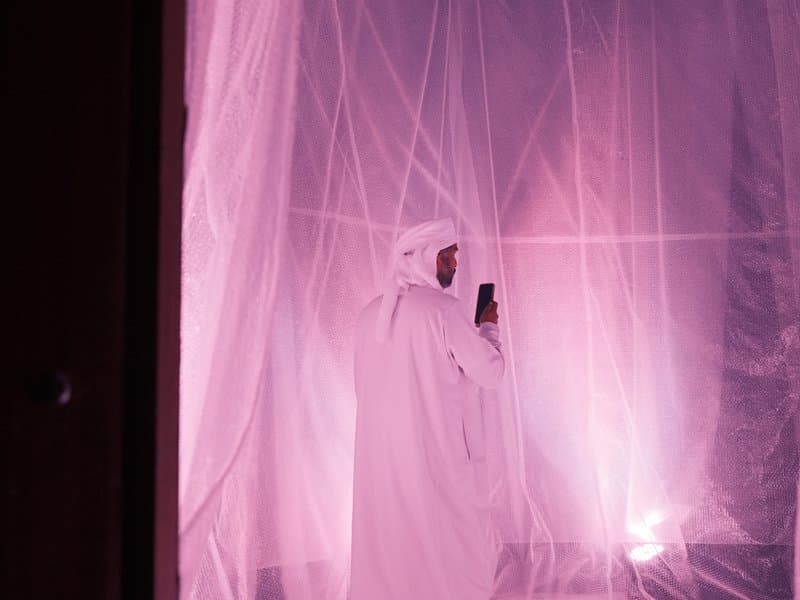 A figure in a white robe stands inside a translucent pink space, illuminated by soft lighting.