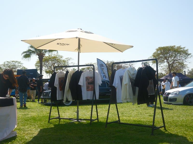 Clothes displayed on racks under a large umbrella at an outdoor market on a sunny day.