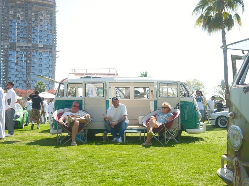 Three people sitting in front of a vintage Volkswagen bus on a sunny day at an outdoor event.