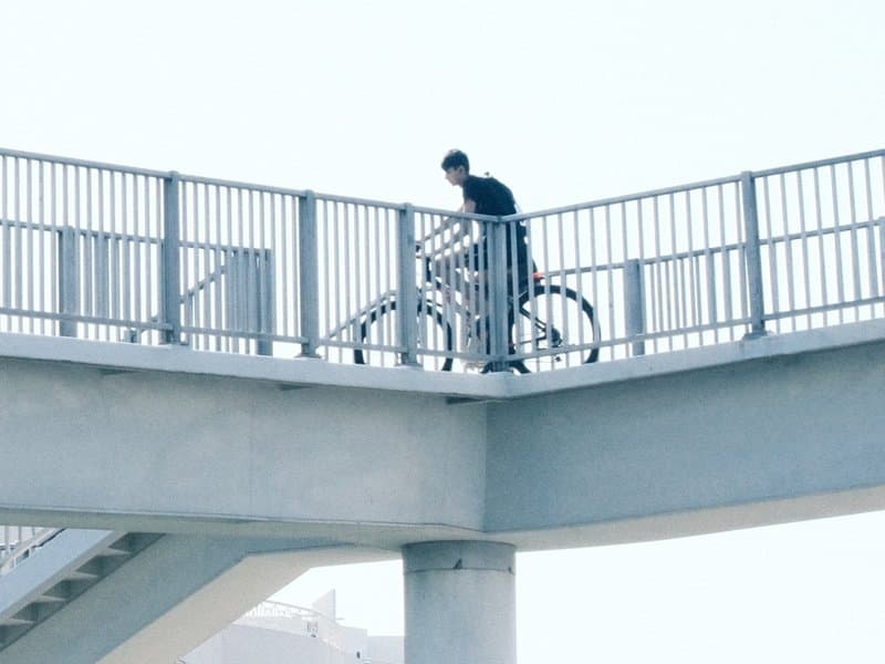 A person walking a bicycle on a bridge with a minimalist urban backdrop.