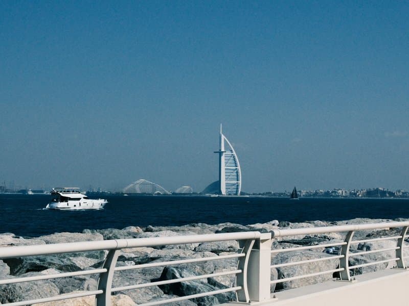 A view of the Burj Al Arab hotel in Dubai with a boat on the water and rocky shoreline in the foreground.