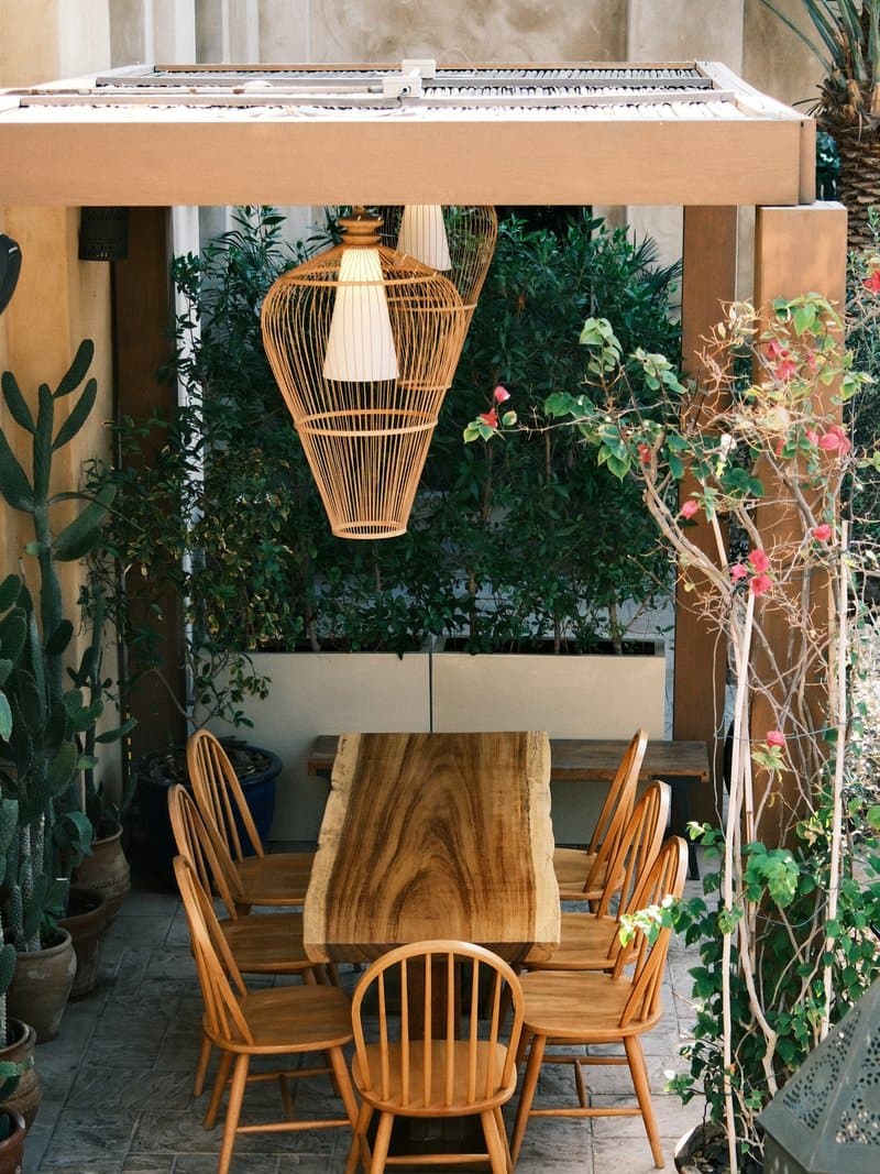 A wooden dining table surrounded by chairs under a pergola with hanging light and greenery.