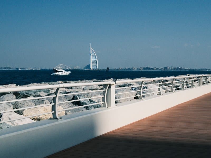 A view of a waterfront promenade with a boat and a tall building in the background against a clear blue sky.