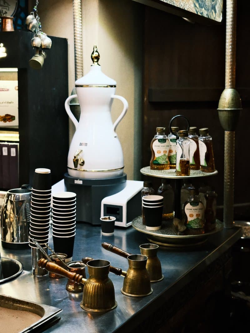 A vintage coffee setup with brass coffee pots, a white coffee urn, and glass jars on a metal counter.