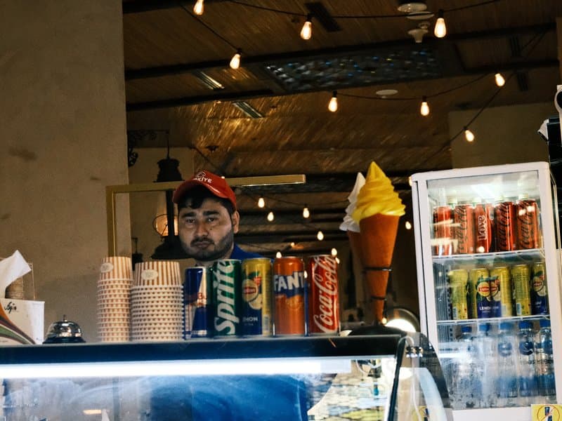 A man behind a counter with soft serve ice cream and various beverage cans in a cozy indoor setting.