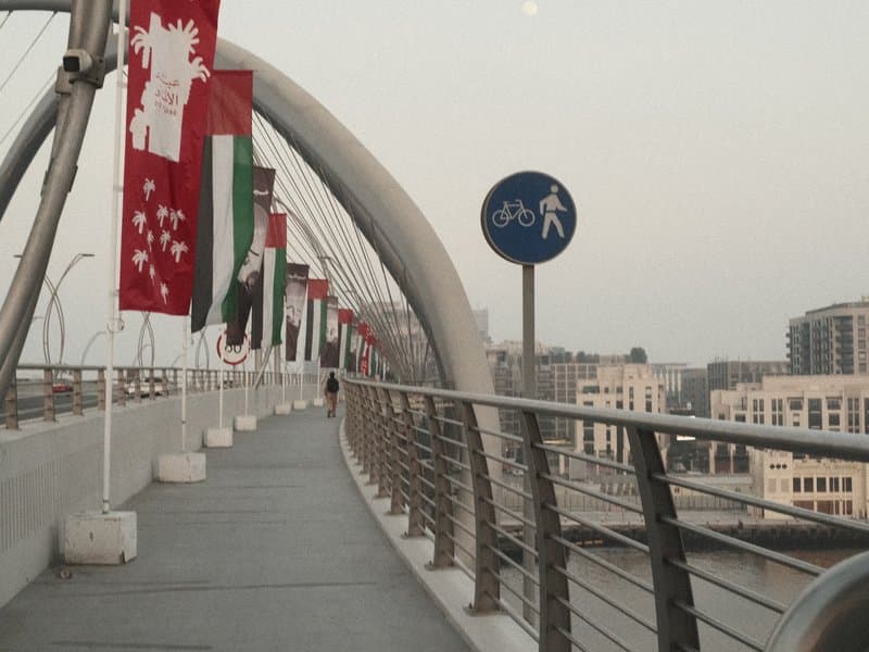 A pedestrian bridge with flags and signage, leading into a cityscape at dusk.