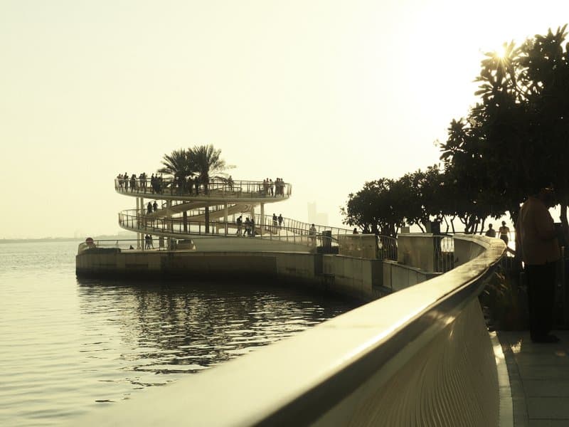 A winding walkway along a waterfront with a spiral structure and silhouetted trees at sunset.