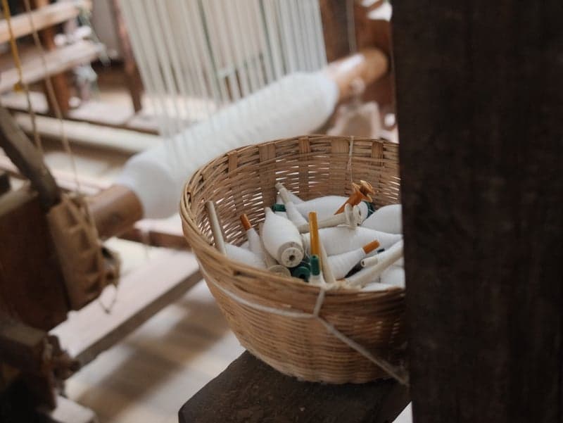 A woven basket filled with spools of thread in a weaving workshop, with looms in the background.