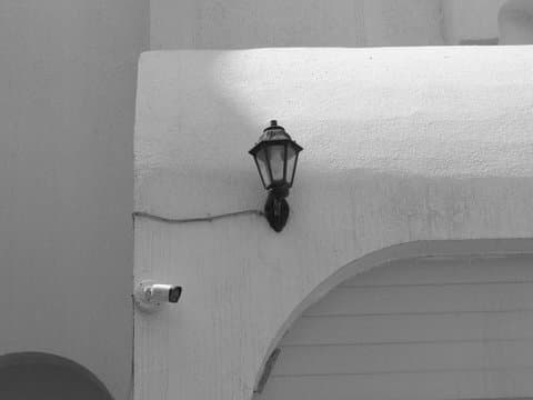 A black and white image of a vintage lamp mounted on a white wall with a security camera nearby.