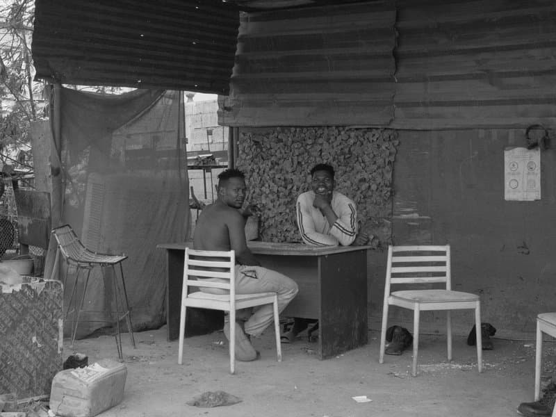 Two men sitting at a table in a rustic outdoor setting with simple chairs and a textured backdrop.