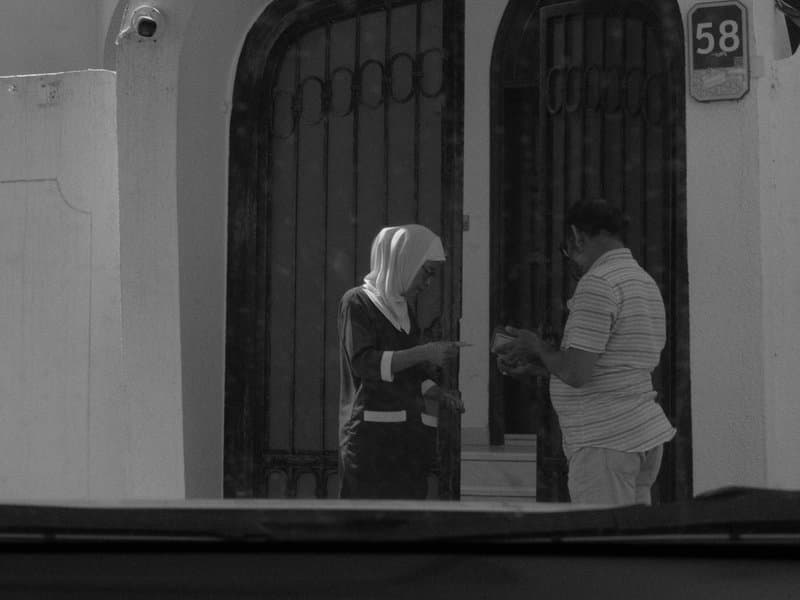 A woman in a hijab and a man exchanging something outside a house with a gated entrance.