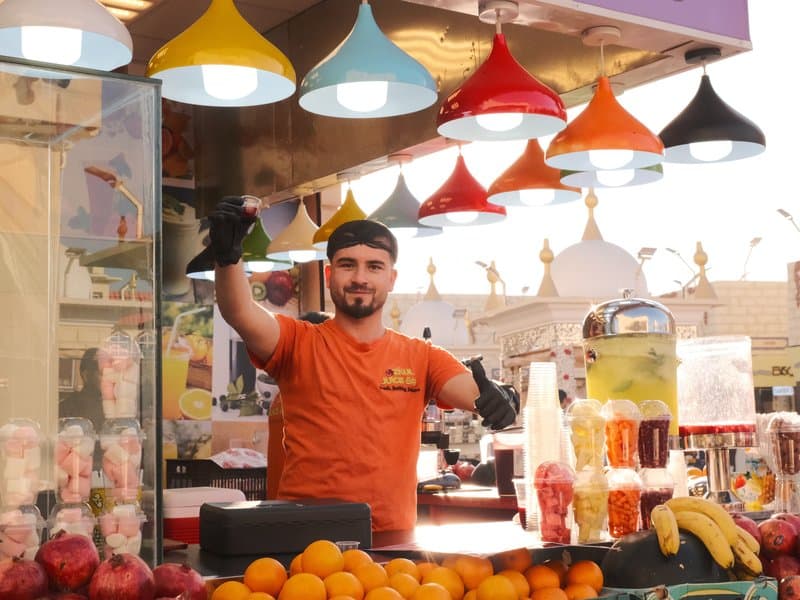 A vendor in an orange shirt stands behind a colorful fruit stall with various fruits and drinks.