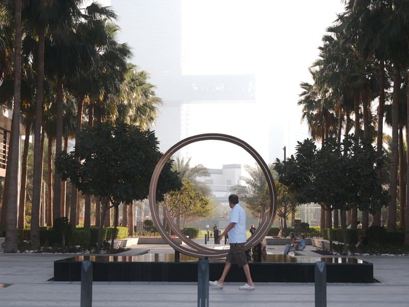 A man walks past a large circular sculpture surrounded by palm trees in an urban setting.