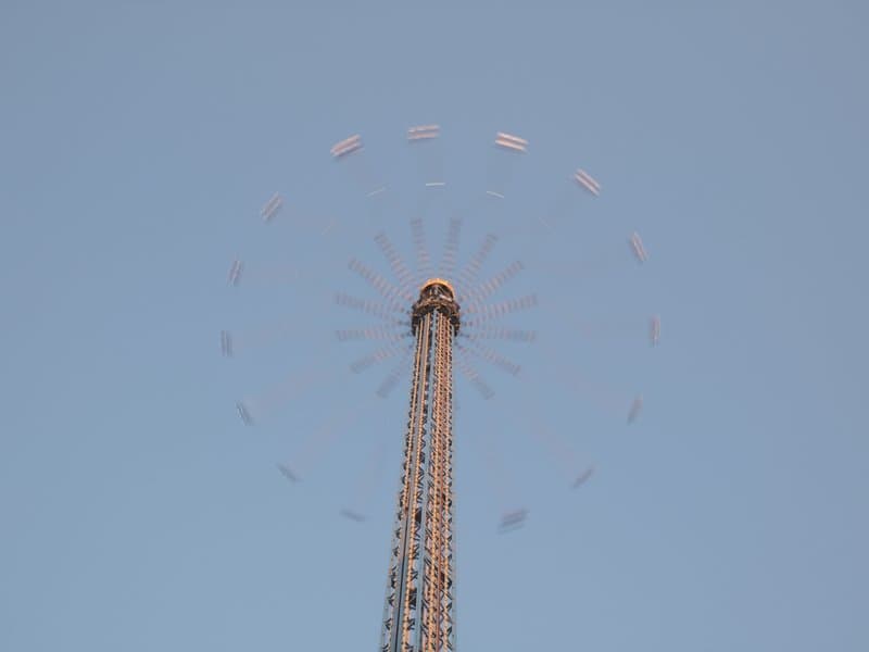 A tall amusement park ride against a clear blue sky, with colorful seats circling at the top.