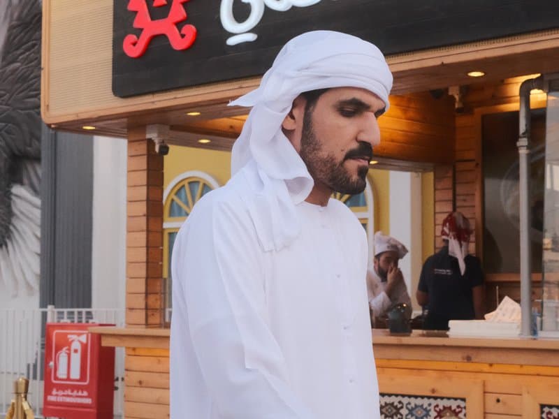 A man in traditional Emirati attire stands in front of a food stall, with another person visible in the background.