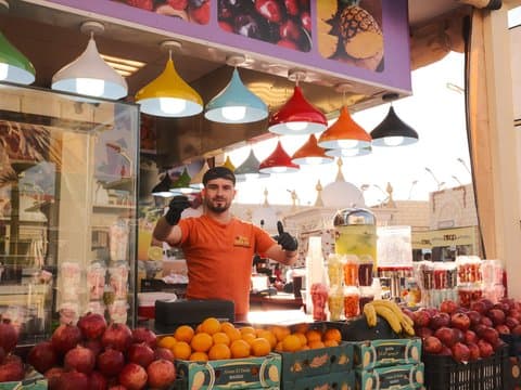 A fruit vendor stands in front of a colorful stall filled with various fruits under bright hanging lights.