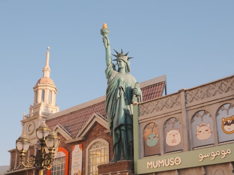 Statue of Liberty replica in an urban setting with colorful storefronts and clear blue sky.