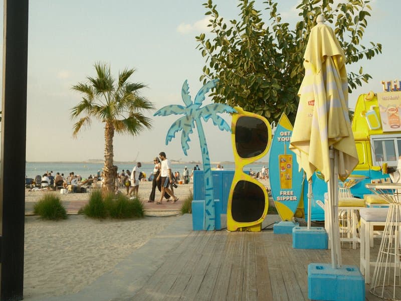 Colorful beach scene with palm trees, oversized sunglasses, and people enjoying the seaside.
