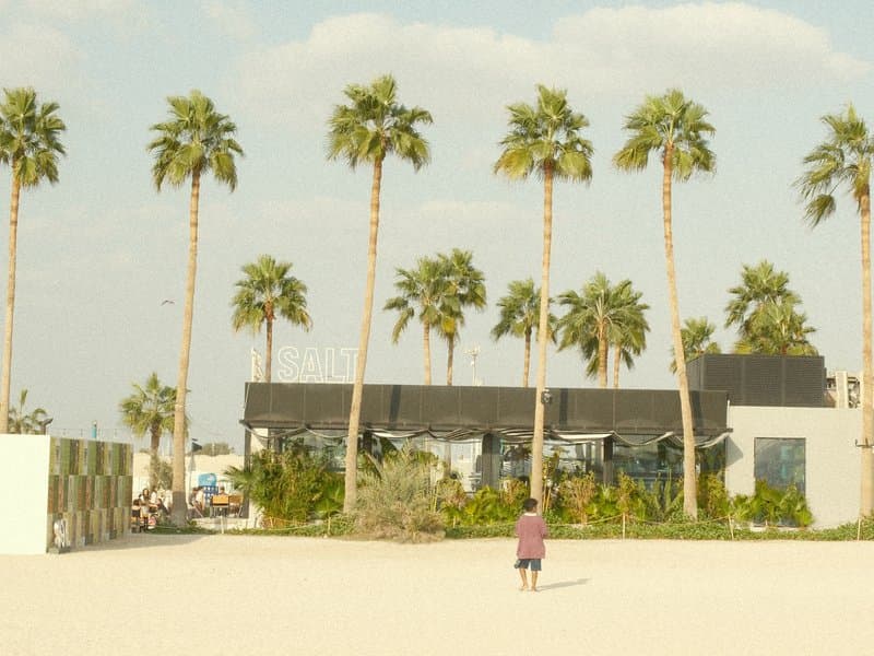 A person walking on a sandy beach with palm trees and a modern building in the background under a bright sky.