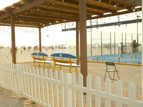 A beachside scene featuring a wooden pergola with colorful benches and a white picket fence.