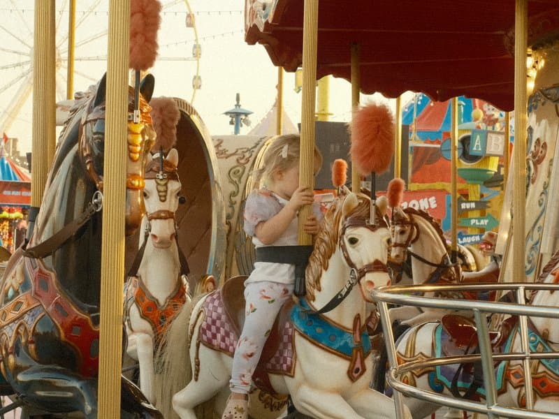 A child riding a carousel horse at a fairground, surrounded by colorful attractions and lights.