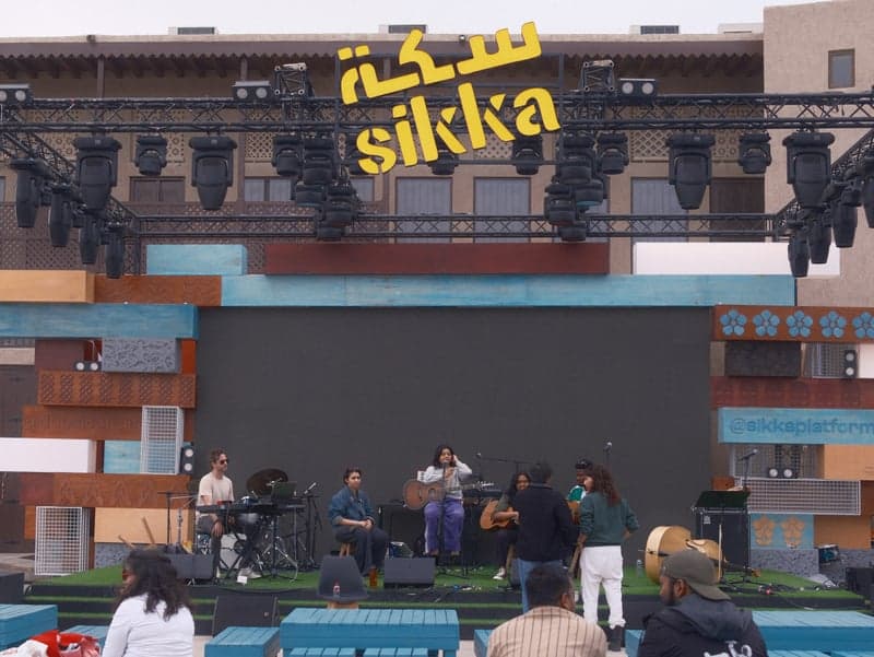 A band performing on stage at an outdoor event with a large screen and colorful backdrop.