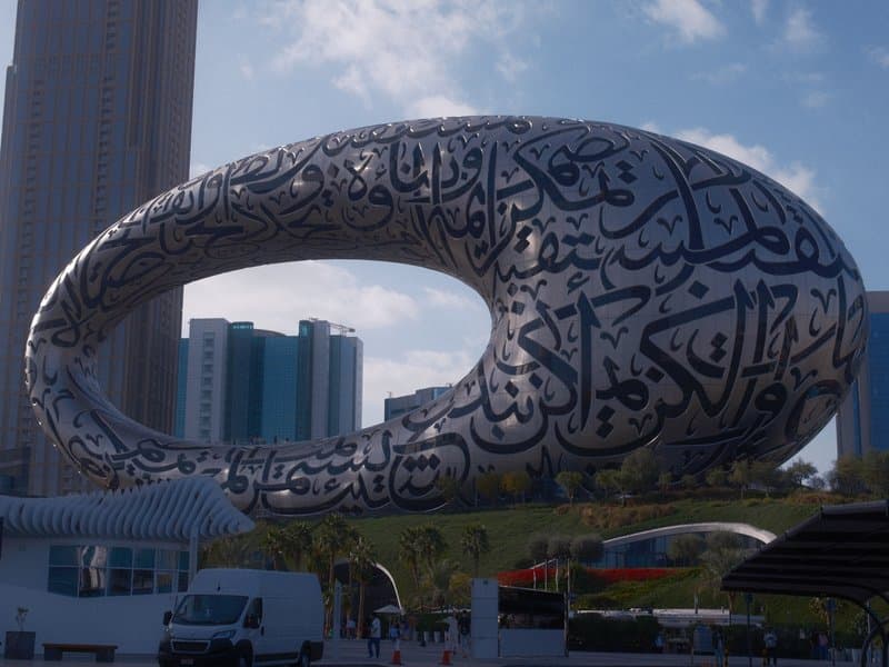 A modern architectural structure resembling an oval shape with Arabic calligraphy, surrounded by buildings.