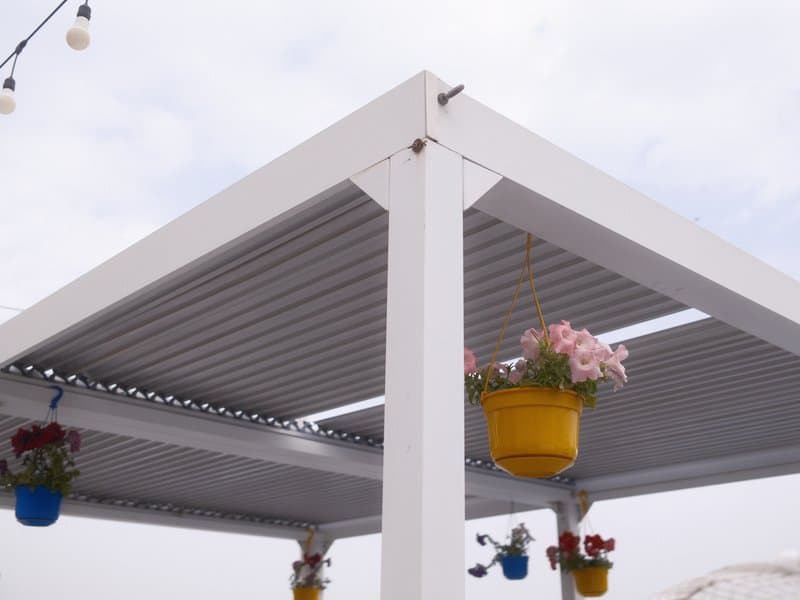 A close-up view of a white pergola with hanging flower pots against a cloudy sky.