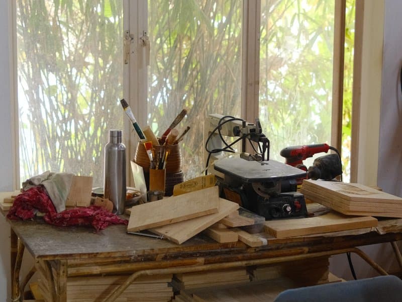 A cluttered workbench with tools, wood pieces, and a backdrop of greenery through a window.