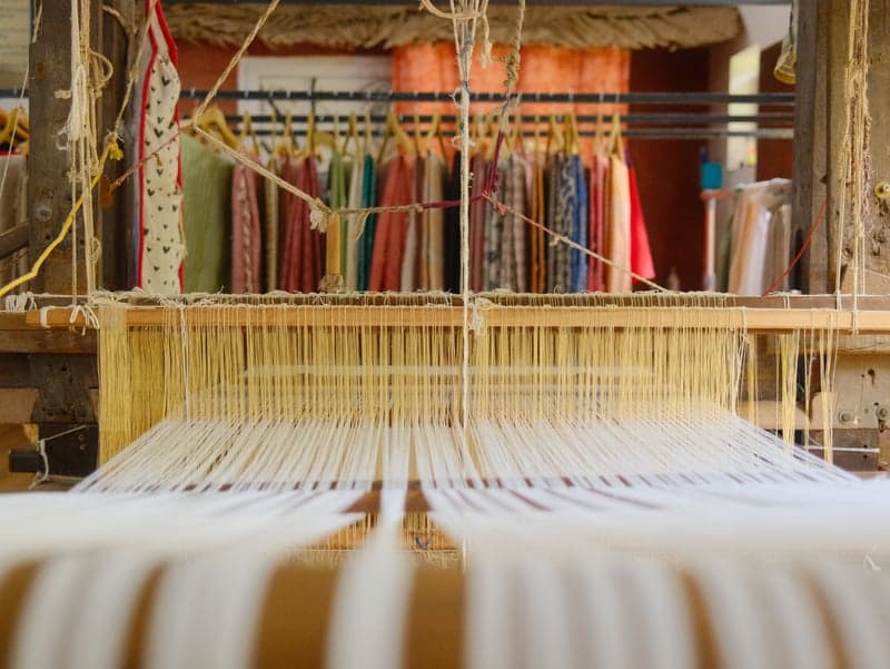 A weaving loom with threads in focus, colorful fabrics hanging in the background.