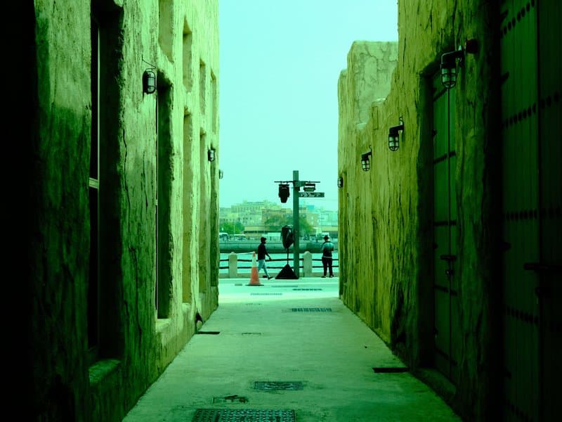 Narrow alleyway with greenish tint leading to a waterfront view, people in the distance.