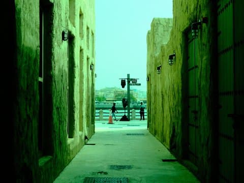 Narrow alleyway with greenish tint leading to a waterfront view, people in the distance.