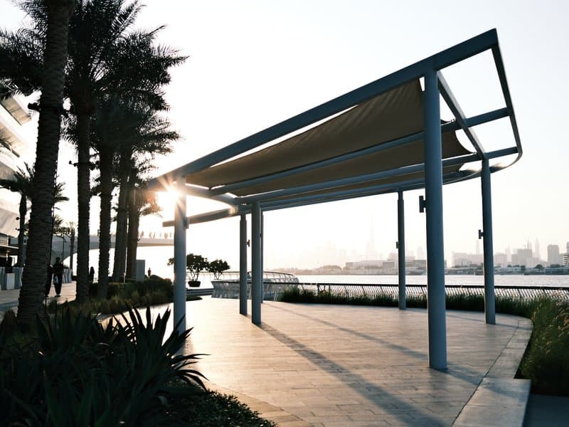 A modern pavilion with a sunshade, surrounded by palm trees and a waterfront view at sunset.