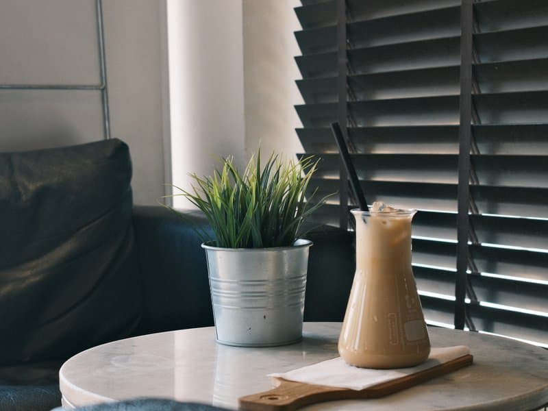 A glass of iced coffee on a marble table next to a small potted plant in a cozy indoor setting.