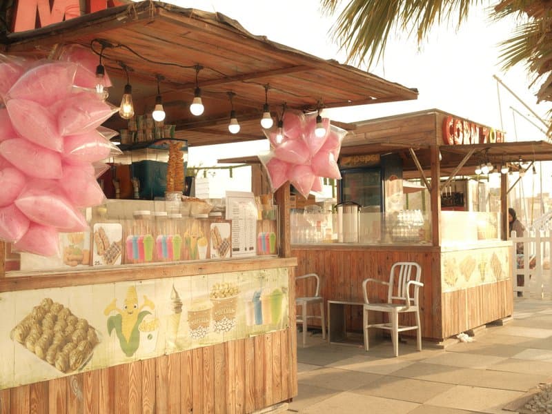Colorful food stalls with pink decorations under warm lighting, set against a coastal backdrop.