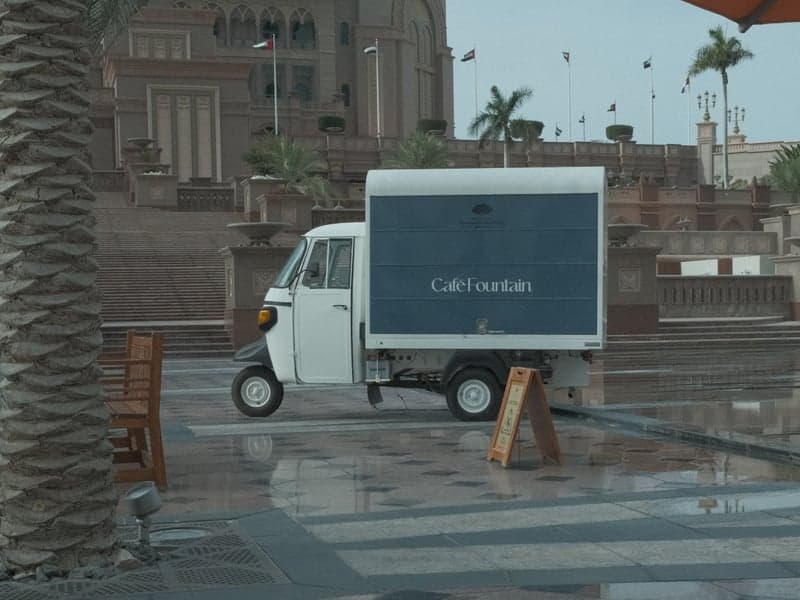 A small white delivery vehicle parked on a wet plaza with palm trees and a large building in the background.