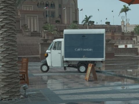 A small white delivery vehicle parked on a wet plaza with palm trees and a large building in the background.
