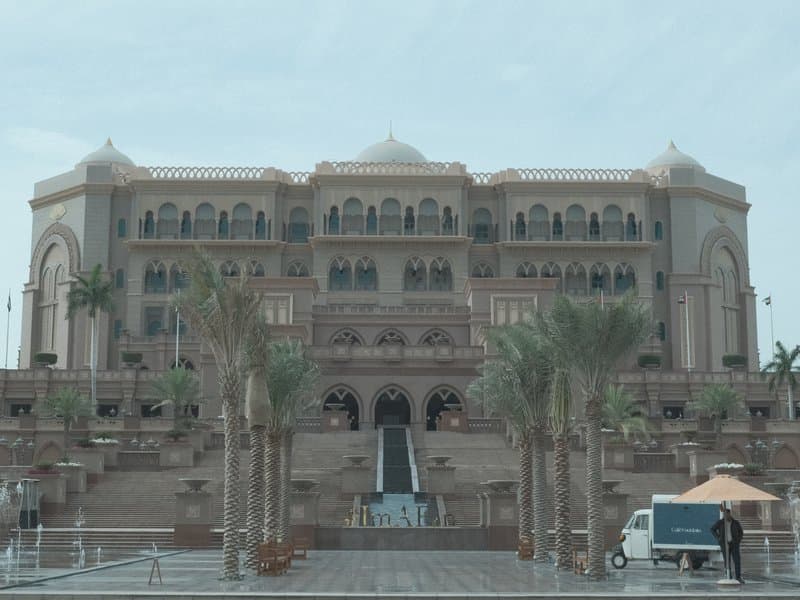 A grand architectural building with palm trees in the foreground and a cloudy sky above.
