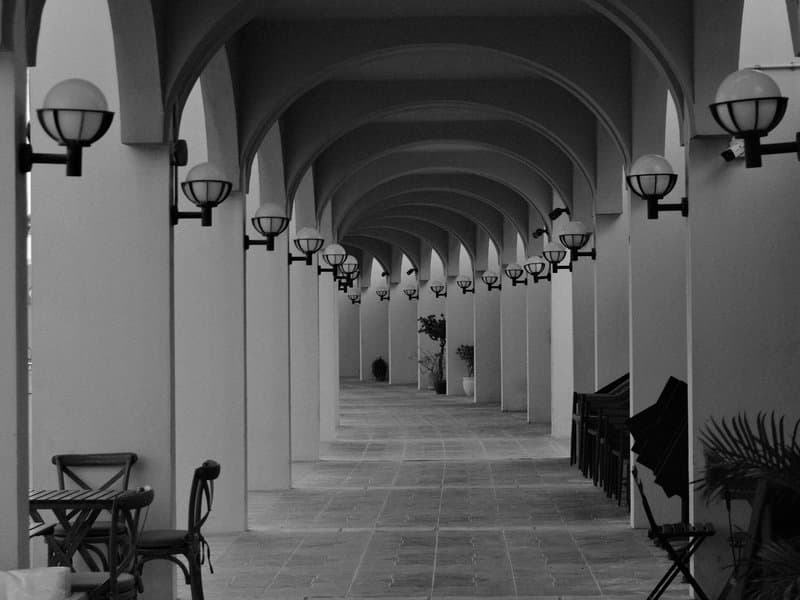 A long corridor with arches and lamps, featuring benches and a tiled floor in black and white.