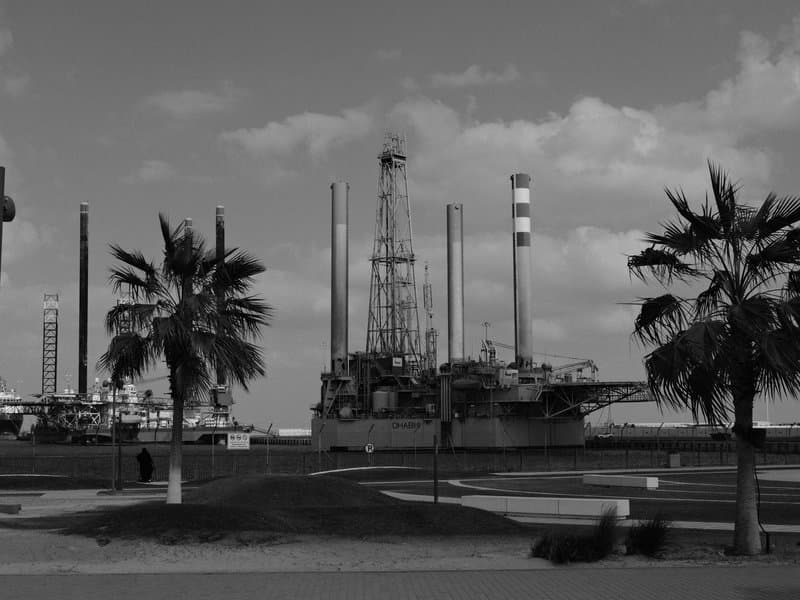 A large oil rig stands prominently against a cloudy sky, surrounded by palm trees.