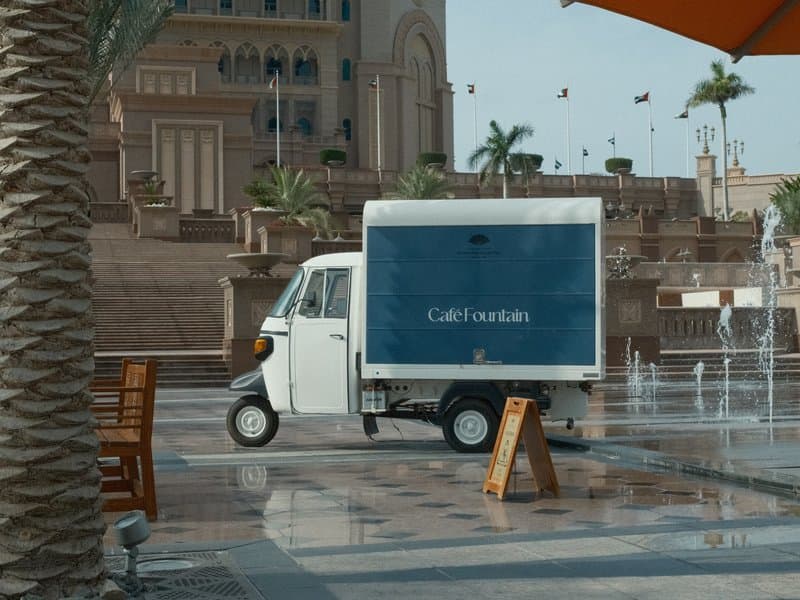 A small delivery truck parked near a fountain in a plaza with palm trees and a large building in the background.