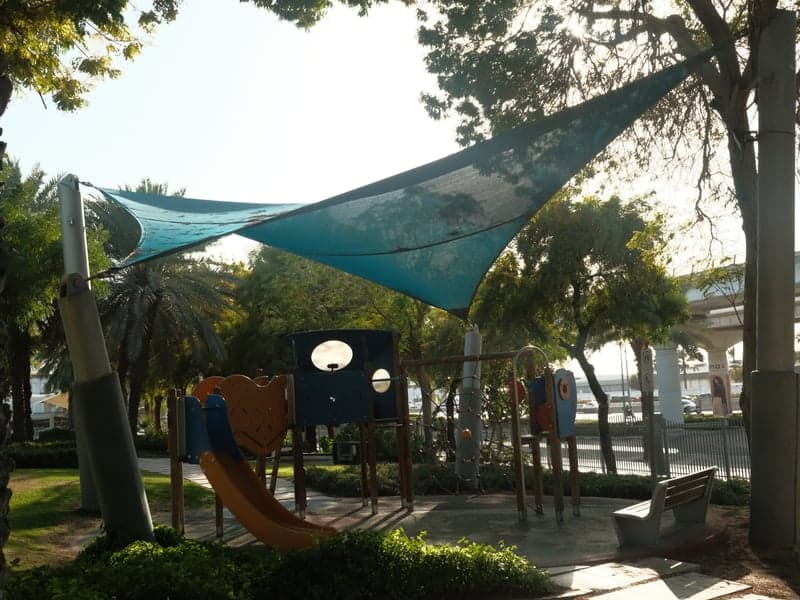 A playground with colorful equipment shaded by a large blue canopy, surrounded by palm trees.