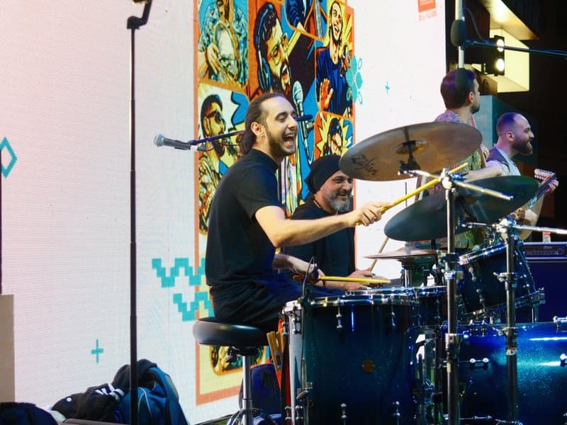 A drummer performing on stage with a colorful backdrop and band members in the background.