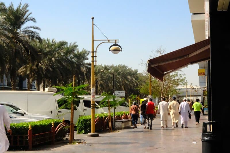 A busy urban street scene with pedestrians walking along a palm-lined sidewalk under a clear blue sky.