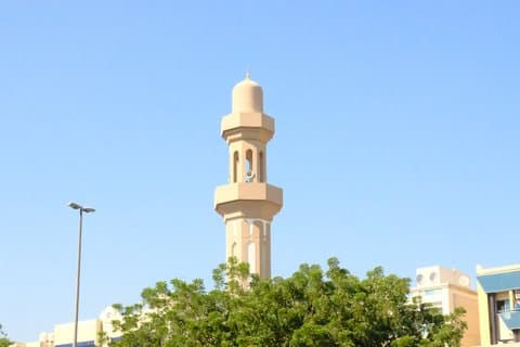A tall minaret rises against a clear blue sky, surrounded by green trees and urban buildings.