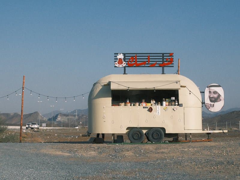 A vintage food trailer with a sign, set against a clear blue sky and distant mountains.