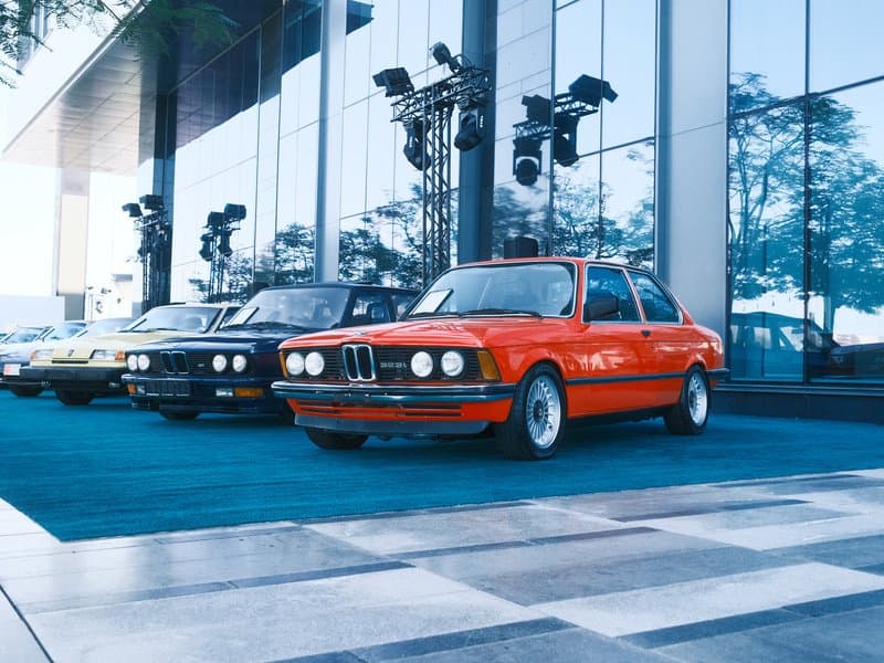 A lineup of classic cars, including a bright orange BMW, in front of a modern glass building.
