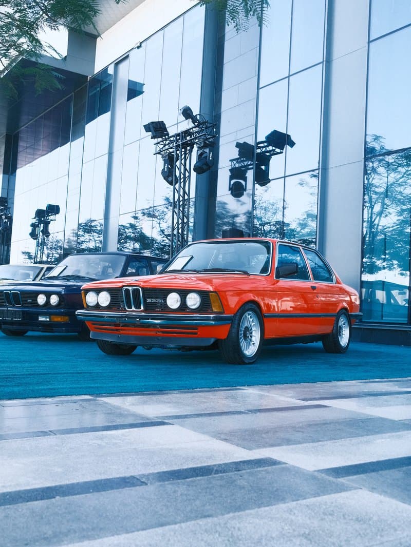A classic orange BMW car parked in front of a modern glass building with another car in the background.